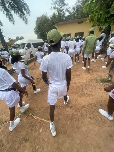 A group of young adults, dressed in white t-shirts, white shorts, and green caps, are gathered outdoors on a dirt path. Many are facing away from the camera, walking in the same direction. A white van, labeled "FETHI" and with a red cross symbol, is parked in the background. The scene appears to be at a camp or training facility, given the uniform attire and the presence of what could be a medical vehicle. The environment is somewhat unkempt, with visible tree roots and scattered debris on the ground. The weather seems overcast. The group's attire suggests they might be participants in a youth service program or a similar organized event. This image captures a moment in a communal gathering, possibly on a field day or during a period of organized activity.