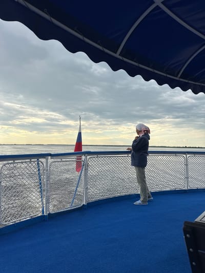 The image shows a person standing on the deck of a boat, looking out at the water. The boat has a blue deck and a white railing. A Russian flag is flying from a pole on the boat. The sky is cloudy. The person is wearing a dark jacket, light-colored pants, and white sandals. They have a light-colored headscarf or hat on. The location appears to be a body of water, possibly a river or lake, with a distant shoreline visible. The scene suggests a calm day, with the person enjoying the view from the boat. There is no specific indication of the city of Zuevo, Russia, in the image itself, but the presence of the Russian flag implies a Russian context.