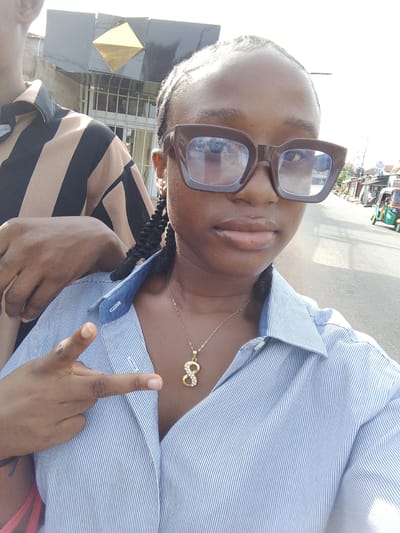 A close-up selfie-style outdoor photograph captures two individuals on a street in Ohokobe Ndume, Nigeria, under bright daylight. The primary subject, a person with dark skin, occupies the foreground, wearing large, dark-framed rectangular eyeglasses that obscure most of their eyes. Their lips, with a slight sheen, are visible, and their hair appears to be styled in braids. They are dressed in a light blue and white pinstriped collared button-up shirt, partially unbuttoned, revealing a gold chain with a diamond-studded pendant resembling the number '8' or an infinity symbol. Their left hand is raised, forming a "peace" or "V" sign with the index and middle fingers.

To the left of the main subject, another person is partially visible, clad in a light brown and black striped short-sleeved collared shirt. Their arm rests casually on the main subject's shoulder.

The background reveals a sunlit street scene characteristic of a local urban or suburban area. Buildings line the paved road; one prominent dark-colored building directly behind the partial subject features a golden, diamond-shaped architectural embellishment. Further down the street on the right, a green and yellow three-wheeled auto-rickshaw (tuk-tuk) is visible, alongside other indistinct structures. The bright, direct sunlight suggests a clear, sunny day, likely late morning or afternoon. The overall impression is casual and relaxed. No discernible text is visible on signs or clothing.