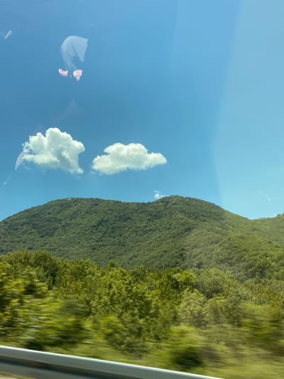 The image depicts a scenic view of a lush green mountain under a clear blue sky with scattered white clouds. The foreground shows a blurred line of trees, suggesting movement, possibly from a vehicle. In the upper left corner, a reflection shows a person's legs and feet. The image appears to be taken from inside a vehicle looking out at the landscape. The location is specified as Vrela, Montenegro.