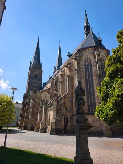A long-shot, eye-level, exterior view captures a stone church with multiple spires and tall, arched windows under a bright blue sky with scattered clouds. The church architecture suggests a Gothic style, with detailed stone carvings and buttresses. In the foreground, a weathered stone statue stands on a pedestal, partially obscured by a lush green tree. The ground is paved with cobblestones, and a lamppost stands to the left of the frame, with a grassy area in the foreground. Another building with a light-colored facade and green shutters is visible in the background, to the left of the church. The scene is illuminated by clear daylight, suggesting it is daytime. The overall atmosphere is peaceful and serene. No text is visible in the image.