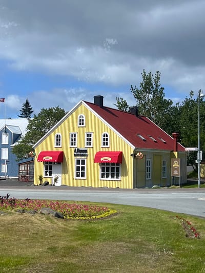 A yellow building with a red roof and red awnings stands on a corner lot in Hafnarfjörður, Iceland. The building is a restaurant called "A-Mensa Grill." The building has several windows and a large door with a red awning above it. There is a sign that says "Grill" on the side of the building, and a sign that says "A-Mensa" over the door. There are some trees in the background, and a small flower garden in the foreground. The street is paved and has a small traffic island with a flower bed. The sky is cloudy and blue. The scene is taken at 64.07 latitude and -21.958 longitude.