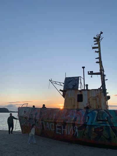 This image depicts a beached, derelict ship on a sandy shore in Teriberka, Russia, during sunset. The ship, heavily vandalized with colorful graffiti, has a rusty and weathered appearance. The sky displays a gradient of blue to pale orange, indicative of twilight. In the foreground, a man in camouflage clothing is standing near the ship, holding a rope. A young girl in casual attire is walking away from the ship towards the viewer. In the background, a few other silhouetted figures can be seen near or on the ship. The sea is calm and to the left of the ship, with a distant coastline visible. The overall scene conveys a sense of abandonment and decay against a backdrop of natural beauty.