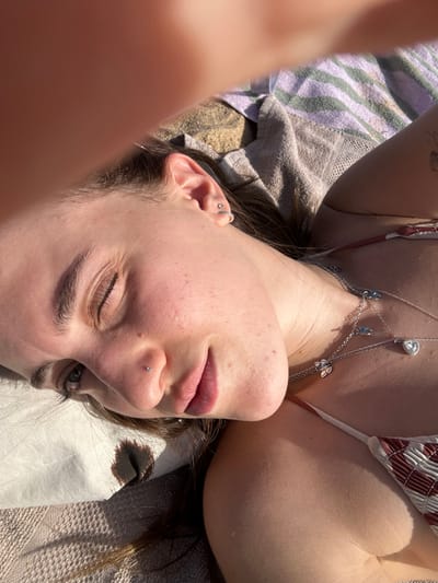A woman lies on her back, eyes closed, wearing a bikini top and a silver body chain. Her face is freckled, and she has piercings in her ear and nose. She rests on a striped towel that partially obscures the beach sand. The sunlight casts shadows and illuminates her skin. A hand is partially visible in the lower left corner.