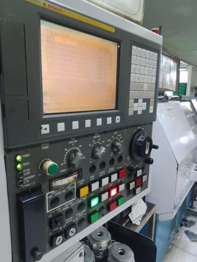 The image shows the control panel of a CNC machine in an industrial setting. The panel features a screen displaying numerical data, a keyboard with alphanumeric keys, various knobs, buttons, and indicator lights. The screen displays "OFFSET GEOMETRY" and other technical information. Prominent labels include "FANUC," "POWER," "READY," "SPINDLE SPEED," and "SPINDLE LOAD." There are also buttons for functions like "CYCLE MODE," "PROGRAM PROTECT," and "COOLANT." The machine appears to be in operation or ready for operation, with some indicator lights illuminated. In the background, parts of other industrial machinery and the general environment of a factory or workshop are visible. The overall scene depicts a functional piece of industrial equipment. The specific location in the city of Ciantra, Indonesia, is not evident from the image itself but is provided as context.