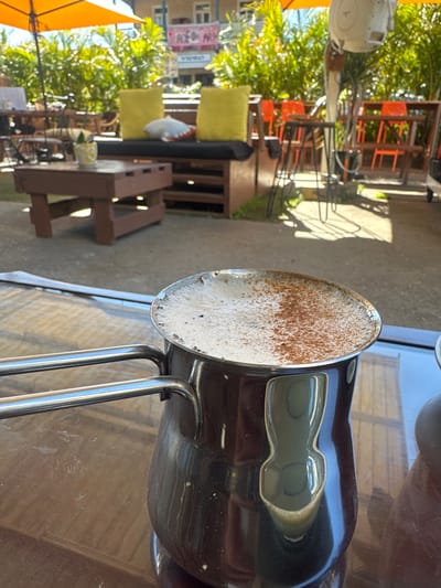 The image shows a close-up of a coffee pot with frothy coffee topped with cinnamon, resting on a wooden surface. In the background, an outdoor seating area is visible, with tables, chairs, and lush green foliage under an orange umbrella. The scene appears to be an outdoor cafe. The presence of banners with text that appear to be in English, such as "ACE OF ARC" and "VEGAS", along with the style of the outdoor seating and vegetation, suggests a casual, possibly tropical or subtropical setting. The general atmosphere is relaxed and sunny. Given the question about the location in Bellingen, Australia, this image likely depicts a cafe or restaurant in that area, known for its natural beauty and outdoor dining options.