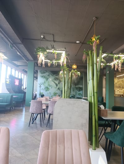 The image shows the interior of a restaurant. The restaurant has a modern design with green and pink seating. There are bamboo-like decorations and artificial plants throughout the space. In the background, a large window offers a view of the city. A neon sign that reads "SALAT" is visible on the left side of the image. The floor is tiled in a marble pattern. There are no people visible in the image.