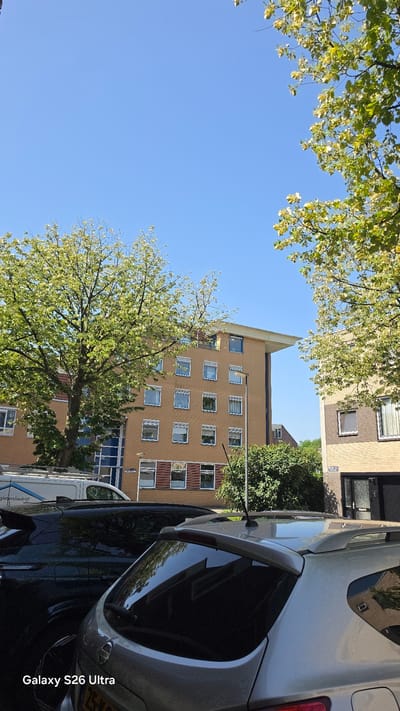 This media file depicts an outdoor scene on a sunny day. The primary subjects are a modern apartment building, trees with lush green foliage, and parked cars. The apartment building is tall and appears to be a light brown or tan color, with multiple stories and numerous square windows. The trees are in full bloom, with light green leaves and some white blossoms, suggesting it is spring or early summer.
The image is oriented vertically and appears to have been taken from a low angle, looking upwards. The clear blue sky indicates fair weather. Several cars are parked along what appears to be a street or residential area in front of the building. One car, a light-colored hatchback, is prominently featured in the foreground on the right side of the image, with its rear visible. Another vehicle, possibly a van, is partially visible to the left of the hatchback.
There are no people visible in the scene. The overall atmosphere is peaceful and ordinary, suggesting a typical day in a residential urban or suburban environment. The location context provided is Vlaardingen, Netherlands, which aligns with the architectural style of the buildings and the type of cars.
A notable detail is the text "Galaxy S26 Ultra" printed vertically on the right side of the image, likely indicating the device used to capture the photograph. There is also some faint text visible on the side of the white van on the left, but it is too blurred to be legible. The time of day appears to be daytime, given the bright sunlight casting shadows and illuminating the scene.