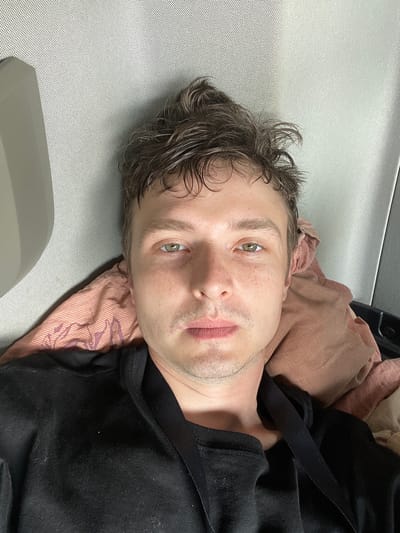The image is a selfie of a person lying down, with their head resting on a pillow. The background appears to be the interior of a vehicle, possibly a truck or a car, with grey textured material on the left and right. The person has short, messy brown hair and is wearing a black hooded sweatshirt. Their eyes are open and looking directly at the camera. The lighting is soft and natural. There are no discernible landmarks or specific features that would indicate a location in Edewecht, Germany or any other specific city.