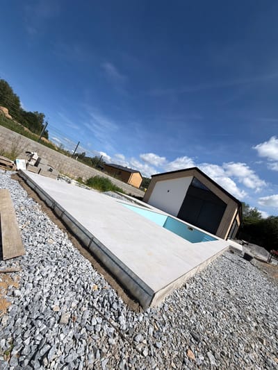 The image captures a construction site in Horní Planá, Czechia, focused on the unfinished area around a swimming pool. A white, tiled surface, presumably the pool deck, covers a significant portion of the foreground, with a section of bright blue water visible within a recessed pool area. The deck is bordered by a metal formwork system and sits atop a foundation of gravel and sand. In the background, a modern house with a dark, pitched roof and a white facade is situated on an incline. To the left, a retaining wall constructed of concrete blocks and topped with a metal fence marks the edge of the property. The scene is set under a clear blue sky with scattered white clouds, suggesting fair weather. The overall impression is one of ongoing construction, with materials and equipment hinting at the development of residential property.