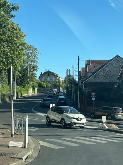 The image depicts a street scene in L'Haÿ-les-Roses, France, under a clear blue sky. A white Renault Captur is stopped at a pedestrian crossing, with its front license plate reading "DP 209 FXN". To the right of the Captur, a darker colored SUV, possibly a Peugeot 5008, is also present. In the background, houses with tiled roofs line the street, and lush green trees flank the road. A "30" speed limit sign is visible on a pole. Traffic appears to be moving slowly uphill, with several other vehicles in queue. A traffic light is situated on the left side of the road. The overall scene suggests a typical day in a residential area with moderate traffic.