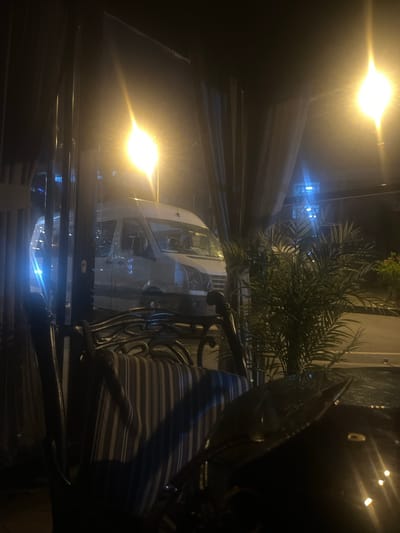 The image depicts a night scene viewed through a window, likely from inside a covered outdoor seating area of a cafe or restaurant. In the foreground, a striped cushion is visible on a metal chair. Beyond the chair, a white van is parked on a street. A street light with a bright, diffused glow illuminates the scene from the upper right. To the right of the van, a palm plant is situated in a pot. The overall lighting is dim, with prominent highlights from artificial light sources. The location is specified as being in the city of Bayrampaşa, Turkey.