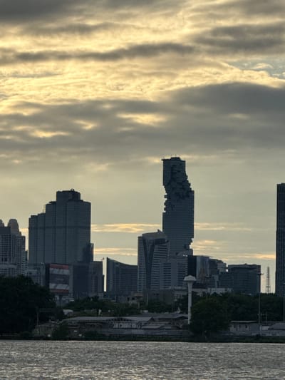 This is a full shot, taken from a body of water, of the Bang Kachao Subdistrict in Thailand. The sky is filled with a mix of dark grey and light yellow clouds. In the foreground, the water is rippled, indicating movement. Dominating the background is a cityscape with several tall buildings, the most distinctive being a uniquely shaped skyscraper with a jagged, pixelated appearance. The buildings are silhouetted against the cloudy sky. Below the skyline, there are more structures and dense green trees. The overall scene suggests an urban environment with a body of water, likely a river or canal, in the foreground, and a cloudy sky above.
