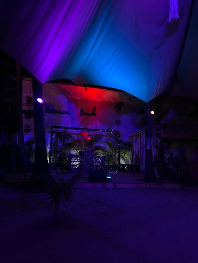 The image depicts a nighttime scene in Santa Teresa, Costa Rica, at the location specified by the coordinates 9.632 latitude and -85.158 longitude. The scene is illuminated by purple and blue lighting emanating from a large, white canopy tent above a stage. A person is positioned behind a table at the stage, facing the audience, with a microphone visible. The stage is made of wood and a small speaker is in front of it. There are palm trees, potted plants, and other vegetation behind the stage. The sign "Banana Beach" is visible in the background above the stage. The scene is dimly lit and the atmosphere appears to be relaxed.