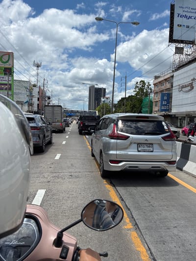 The image shows a busy street scene in Nonthaburi City Municipality, Thailand. A person is riding a scooter in traffic, as evidenced by the scooter's handlebars, mirror, and front fairing visible in the foreground. Other vehicles on the road include cars, a truck, and a Mitsubishi pickup truck. Buildings line the street, with signs in Thai script. The sky is blue with scattered white clouds, indicating a sunny day. The traffic appears to be moderate, with vehicles moving slowly.