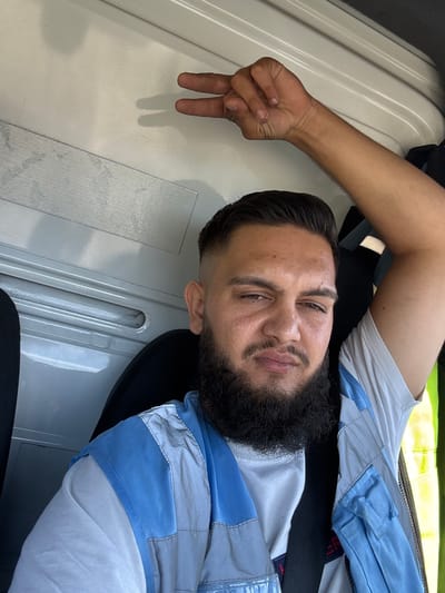 A man is sitting inside a vehicle, possibly a truck, making a peace sign with his hand. He is wearing a light-colored t-shirt and a blue and grey vest. The interior of the vehicle is white and grey. The man has a beard and short hair. The lighting suggests it is daytime. There are no specific visual cues to identify the location as Malmedy, Belgium.