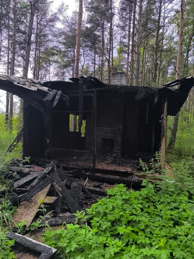 The image depicts the aftermath of a fire at a structure located in a wooded area in Laagri alevik, Estonia. The building, which appears to have been a small house or cabin, is extensively damaged and charred, with only the brick chimney and parts of the walls and roof remaining. Debris, including burnt wooden planks and other structural materials, is scattered around the structure. The surrounding environment is a forest with tall trees and dense green undergrowth, indicating the structure is situated in a natural, possibly rural, setting. There are no people visible in the image. The scene suggests a recent fire that has caused significant destruction to the building.