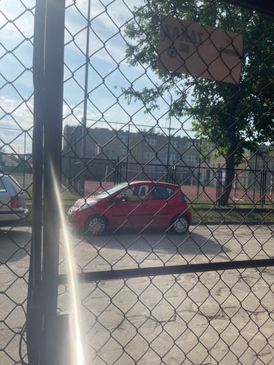 In Konstantynów Łódzki, Poland, through a chain-link fence, a red hatchback car is parked on a paved surface. The car is positioned on the right side of the frame, facing left. Behind the car, a fenced-off outdoor court area with red surfacing is visible, possibly a tennis or basketball court. A building with multiple windows stands in the background, suggesting an urban or semi-urban setting. The sky is partly cloudy. A sign is attached to the fence in the upper right quadrant, but its text is unreadable. A bright lens flare emanates from the left side of the image, partially obscuring the view. Another silver car is partially visible on the left edge of the frame.