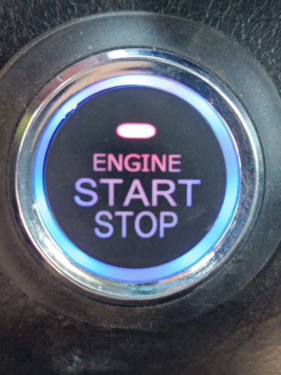 The image shows a close-up of a car's push-button start. The button is circular with a metallic rim and a glowing blue light around its circumference. In the center of the button, the word "ENGINE" is displayed in pink text above "START" and "STOP" in purple text. A pink indicator light is visible above the word "ENGINE". The button is surrounded by a textured black surface, likely part of the car's dashboard. There are no people visible in the image. The location in the city of Saran, Kazakhstan, is not discernible from the image itself.