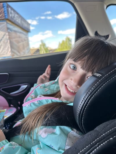 A young girl with blue eyes and light brown hair is looking out the window of a car, smiling and waving. She is wearing a light blue jacket with a pink zipper and unicorn patterns. The car seat headrest is black leather with white stitching. Outside the window, there is a building with signage that is difficult to read, and a blue sky with some clouds. The image appears to have been taken at an angle. There is no specific evidence to confirm it was taken in Миндерево, Russia.