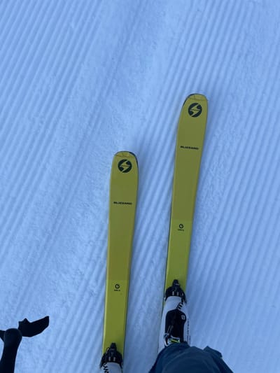 A first-person view depicts a skier's bright yellow Blizzard Zero G skis and Salomon ski boots on a meticulously groomed, ridged snow slope in Vallorcine, France. The skis are parallel, with bindings securing the boots. A partial black ski pole grip is visible in the lower-left, and the skier's dark blue pant leg is seen at the bottom center. The snow is pristine, showing distinct parallel corduroy patterns indicative of recent grooming.