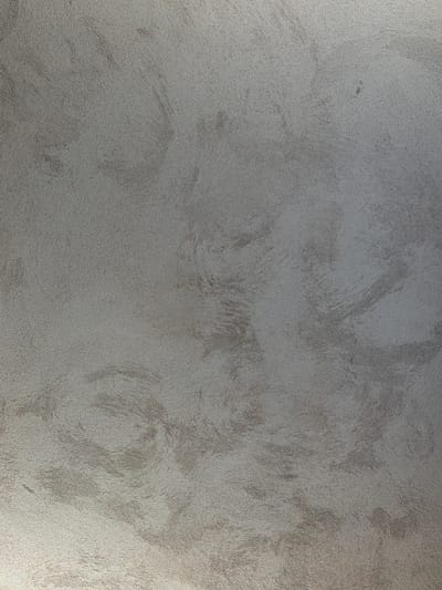 The image is a close-up, full-frame shot of a textured, gray wall with subtle swirling patterns. The texture appears to be a fine-grained plaster or stucco, with variations in sheen suggesting different angles of light catching the surface. There are no people, objects, or discernible environmental features visible within the frame. The lighting is soft and diffuse, creating a slightly gradient effect from the brighter bottom to the darker top. This image does not provide any information about the city of Piacenza, Italy, or any specific location within it.