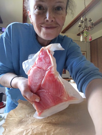 A woman in a blue sweatshirt holds a vacuum-sealed raw tuna steak in a plastic package, presenting it towards the camera. Her right hand supports the steak from underneath, while her left hand holds the package. The tuna is bright pink with visible marbling. The background shows a ceiling with wooden beams, a chandelier, and a wall with a framed picture. The scene appears to be indoors, likely a kitchen or dining area, but its specific location in Alexandrovskaya, Russia, cannot be determined from the visual information provided.