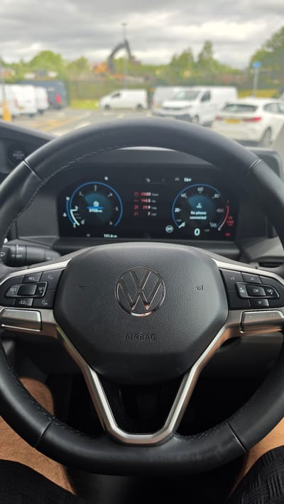 The image captures a close-up view of the interior of a Volkswagen vehicle, focusing on the steering wheel and dashboard. The steering wheel, made of black leather with silver accents, features the Volkswagen logo prominently in its center, along with various control buttons. The dashboard displays a digital screen with information, including speed, mileage, and system alerts such as "No phone connected." The "AIRBAG" label is visible below the steering wheel.
Outside the vehicle, visible through the windshield, is a street scene. The environment appears to be an urban or suburban area, with multiple white vans and other vehicles on the road. In the background, a construction site is evident with an excavator. The sky is overcast.
While the image clearly depicts the interior of a vehicle and a street scene, there are no specific visual cues within the image itself that definitively place the location in Bolton, United Kingdom. The presence of construction activity and various types of vehicles is common in many urban environments.