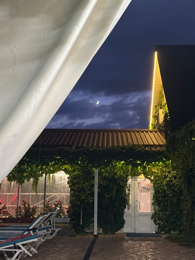 The image depicts a nighttime scene in Yaroslavskaya, Russia, likely an outdoor seating area or resort. In the foreground, a large, white canvas drapes across the left side of the frame, partially obscuring the view. Below the canvas, a corrugated metal roof is visible, covered in lush green vines. A pathway paved with interlocking bricks leads towards a white door framed by more dense green foliage. The door has a silver handle and a frosted glass panel with some signage visible. Red flowers in planters line the area to the left of the pathway, alongside several white lounge chairs with blue and white striped cushions. In the upper portion of the image, the night sky is a deep blue, with scattered clouds and a crescent moon. A modern building with a dark facade and illuminated yellow lighting along its edge is visible on the right. The overall impression is one of a tranquil, dimly lit outdoor space at dusk or night.