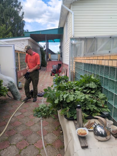 A man wearing a camouflage cap, an orange t-shirt, and camouflage pants is standing on a patterned patio, putting on black gloves. He is near a large plastic bag filled with greenery, a water bottle, and a hose. In the background, there is a white building with a green roof overhang, a glass greenhouse structure, a wood chipper, and some trees. To the right, a table is set with a thermos, a bowl of nuts, a plate of bread, a metal teapot, and two mugs. Oak branches are piled on a tray next to the greenhouse. The scene appears to be a backyard in Velikovo, Russia.
