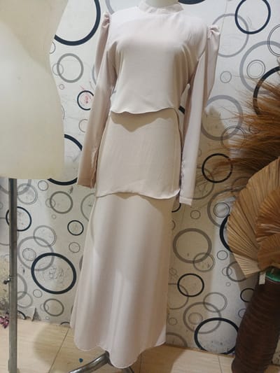 A light-colored, two-piece women's garment, possibly a dress or a skirt and blouse set, is displayed on a mannequin against a patterned wallpaper. The garment has long sleeves and a high neckline. The wallpaper features a repeating pattern of circles in varying sizes and shades of gray and black. To the left of the mannequin is a white, torso-shaped display form. To the right, part of a large brown, ribbed vase is visible, containing dried floral arrangements that appear to be palm fronds. The floor is tiled with a light-colored, geometric pattern. The scene appears to be an indoor setting, possibly a boutique or a display area. There is no indication of the specific location within Payacut, Indonesia.