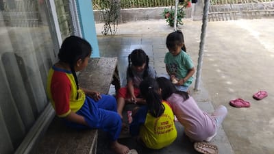 Five young Indonesian girls are gathered in a casual outdoor setting on a concrete patio in Kecamatan Panjalu. One girl, with braided hair, sits on a wooden bench to the left, wearing a yellow and red t-shirt and blue pants. She faces inwards towards the others. Four other girls are seated on the ground, forming a semi-circle.

One girl in a grey t-shirt and red shorts is focused downwards, seemingly playing with something on the ground. Next to her, another girl in a yellow t-shirt and blue pants has her back to the camera; text on her t-shirt reads "PENDIDIKAN ANAK USIA DINI H. NURHIDAYAH SUKABUMI." A third girl, wearing a light green t-shirt and light blue shorts, holds a piece of bread or snack in her hands. The fourth girl on the ground wears a pink long-sleeved shirt and light-colored pants, also looking towards the center of the group. Several of the children are barefoot.

The setting appears to be a veranda or porch beside a building, with a window or glass panel visible to the left. In the background, there's a low wall or fence with bamboo-like structures and some green foliage, suggesting a residential or rural environment. Two pairs of pink flip-flops are neatly placed on the concrete floor to the right. The natural daylight indicates it's daytime, and the children appear calm and engrossed in their activity.