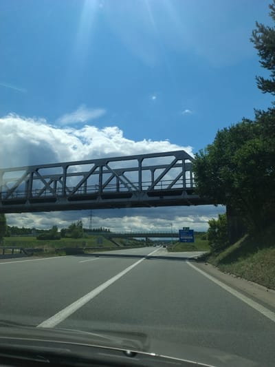 The image is a photograph taken from a vehicle, looking forward along a multi-lane highway. A large, steel truss bridge spans across the highway in the middle distance. The sky is blue with scattered white and dark clouds. On the right side of the highway, there is a grassy embankment with trees. A blue road sign is visible on the right, indicating directions to "PELHRIMOV CHYNOV SEZIMOVOU USTÍ TABOR-MEZICE" and the route number "19". The environment suggests an outdoor, daytime scene on a highway. The specific location is near Tábor, Czechia.