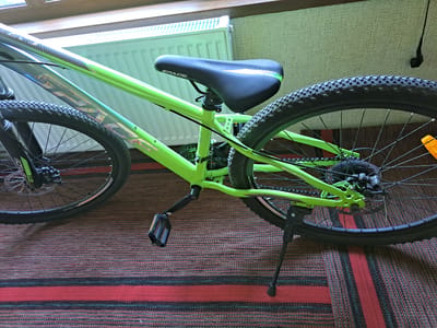 A close-up, eye-level shot captures the rear section of a bright green bicycle with black tires, parked indoors on a red and black striped rug. The bicycle's frame is a vibrant green, with silver and holographic decals visible on the top tube. A black saddle with "SPACE" branding sits atop the seat post. The rear wheel has a knobby tire and a disc brake system. A black kickstand is deployed, holding the bicycle upright. In the background, a white radiator is visible, suggesting the location is a residential interior, possibly in the city of Pervomaisk, Ukraine. Sunlight streams from a window above, creating a bright highlight on the radiator.
