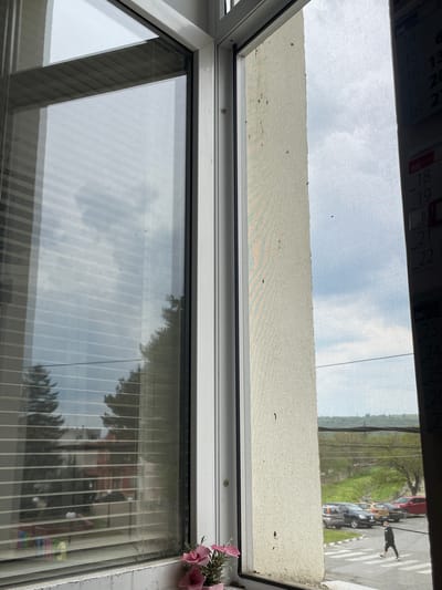 The image depicts a view through a window, obscured by a screen, with a calendar partially visible at the top. The window frame is white, and the scene outside includes a street with vehicles and pedestrians. The calendar shows dates ranging from April 14, 2026 to June 26, 2026. The image does not contain any specific information about the location in Sungurlare, Bulgaria.