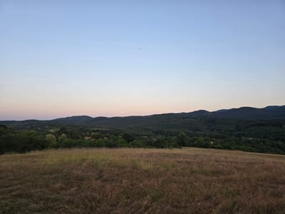 This media file depicts a serene landscape at dusk in Golyama Zhelyazna, Bulgaria. The scene is dominated by a vast, rolling field of dry, tall grass in the foreground, its hues ranging from golden brown to deep purple, suggesting a late summer or early autumn setting. Beyond the field, a dense forest blankets the undulating hills, creating a textured backdrop of dark greens.
Scattered amidst the trees, the roofs of a few houses are visible, hinting at a small rural settlement. The sky above is a gradient of soft blues and pale oranges, transitioning from the clear azure at the zenith to a warmer, dusky tone near the horizon, indicating the approach of evening. A faint, luminous full moon is visible in the sky, positioned above the distant treeline.
In the far distance, silhouetted against the sky, are the majestic shapes of mountain ranges. The overall atmosphere is one of tranquility and peacefulness, evoking a sense of calm as the day concludes. There are no people or specific objects of focus, allowing the natural beauty of the landscape to take center stage. The time of day is clearly evening, characterized by the soft light and the presence of the moon. There is no visible text in the image.