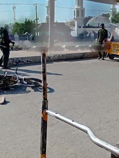 The image captures a street scene in the city of Sani Mai Nagge, Nigeria, under clear daytime conditions. In the immediate foreground, a black and yellow striped metal pole is centrally positioned, with a silver-colored horizontal metal bar extending to the right. On the asphalt road to the left of this pole, a dark-colored motorcycle lies on its left side. A single light-colored sandal rests on the road surface near the fallen motorcycle.
Further back, the paved road is bordered by a raised curb and a low white fence. Two individuals are visible: on the left side of the road, a person dressed in dark clothing walks away from the center of the frame towards the left; their upper body is partially obscured by a dark, amorphous shape. On the right, a male individual stands upright next to a yellow three-wheeled auto-rickshaw. He is wearing a camouflage military uniform and a dark beret, and appears to be observing the scene, facing generally towards the left.
In the background, a large white mosque with multiple domes and minarets is prominent, featuring a crescent symbol atop its main dome. A tall communication tower is visible further in the background against a bright blue sky, along with other structures and trees.