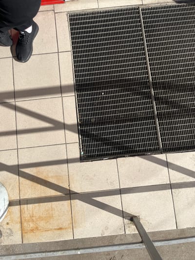 The image shows a close-up view of a person's legs and feet standing on a tiled surface next to a metal grate. The person is wearing black pants and black athletic shoes with white socks. The tiled surface appears to be an outdoor walkway or pavement, with light-colored square tiles. The metal grate is a series of parallel bars forming a grid pattern, likely covering a drain or utility access. Shadows from an unseen structure or object cast angular patterns across the tiles. The scene is located on a street in Apsheronsk, Russia.