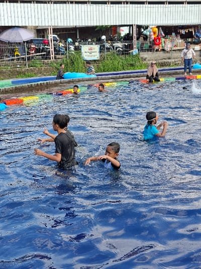 The image shows a swimming pool in Kecamatan Tulung, Indonesia, located at coordinates -7.592 latitude, 110.65 longitude. There are five children swimming in the pool. One child is wearing a black shirt and is swimming on the left side of the image. Another child is wearing a blue shirt and is swimming on the right side of the image. The other three children are swimming in the middle of the pool. There is a blue inflatable raft partially submerged in the water on the left side of the image. The pool is surrounded by a concrete deck and a fence. The scene is taken from a high angle looking down on the pool. The water in the pool is clear and blue, and the sky is blue. The pool is surrounded by lush greenery.