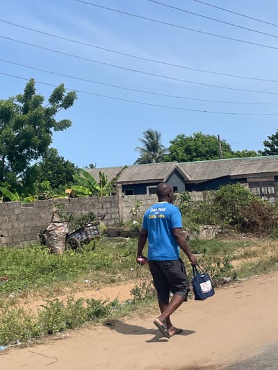The image shows a man walking along a dirt road in Ikoga, Nigeria. He is wearing a blue t-shirt with yellow text that reads "Get Paid with Gab Lotto" on the back and black shorts. He carries a blue bag in his hand and is wearing sandals. The road is bordered by low walls and vegetation. In the background is a building with a corrugated metal roof. Power lines are visible in the sky. A rusted cart leans against a wall.