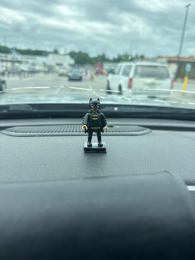 A vertical, close-up shot shows a small Batman figurine mounted on the dashboard of a vehicle. The figurine is black with a yellow bat symbol on its chest, and its arms are bent at the elbows. It is attached to a small, rectangular black base that is adhered to a textured gray surface, likely the dashboard. The dashboard has a circular speaker grille visible to the right of the figurine.
Through the vehicle's windshield, a blurred exterior scene is visible. The scene appears to be a parking lot or street with several vehicles, including a white van in the background. The sky is overcast with clouds, suggesting a daytime scene with potentially poor weather. The overall impression is one of a person driving with a small decorative item on their dashboard. The image's orientation and the nature of the objects suggest the photo was taken from inside the car, looking forward. No visible text is present in the image.