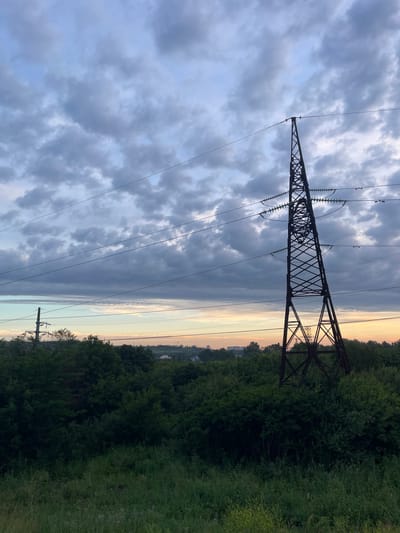 A silhouette of a tall, lattice-style electrical pylon stands on the right side of the frame against a cloudy sky. The pylon is dark brown and its structure is clearly visible, with lines extending from its top and across the sky. Below the pylon and stretching across the lower two-thirds of the image is a dense, dark green canopy of trees and vegetation. In the distance, on the horizon behind the trees and below the sky, a faint line of buildings and more electrical infrastructure can be seen, suggesting an urban or semi-urban environment. The sky is filled with a mix of gray and white clouds, with hints of lighter color towards the horizon, possibly indicating dusk or dawn. The foreground features tall grass. The location is identified as Krymsk, Russia.