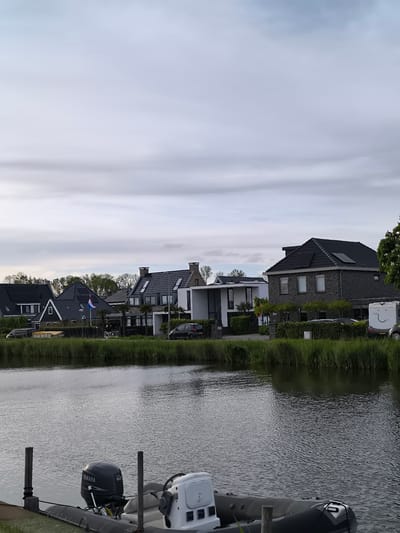 The image shows a view in Oudorp, Netherlands, focusing on a waterfront scene. A body of water, likely a canal or river, occupies the lower half of the frame. A rubber dinghy with a Yamaha outboard motor is docked in the foreground, partially obscuring the water. Pilings protrude from the water near the boat.
Across the water, a row of houses lines the bank. The houses vary in architectural style, with dark-colored roofs and facades. Some have visible solar panels on the roofs. There are several cars and a camper parked near the houses. The grass is lush and green. The Dutch flag is visible on a flagpole in front of one of the houses, next to the street sign "25A".
The sky is overcast with a layer of gray clouds. Overall, the scene depicts a residential area by the water.