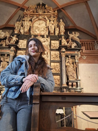 An interior scene captured in the Stiftskirche St. Peter und Alexander (Collegiate Church of St. Peter and Alexander) in Aschaffenburg, Germany, displays a young woman posing in the foreground against a backdrop of a highly ornate Baroque high altar.
**Person:** A single individual, a young woman, is positioned slightly right of the frame's center. She possesses long, wavy, dark brown hair with visible reddish undertones, extending past her shoulders. Her face is turned slightly upwards and to the viewer's left, exhibiting a subtle smile. Her eyes are dark, and her eyebrows are defined. Her hands are clasped and rest on the top edge of a dark wooden structure. She wears a silver-toned ring on the ring finger of her left hand and a similar ring on the middle finger of her right hand. Her fingernails are manicured with a light polish.
**Attire:** The woman is dressed in a light-blue denim jacket, which features a white, sherpa-like fleece lining visible at the collar and cuffs. The jacket has silver-toned buttons and breast pockets. A dark garment, possibly green or black, is visible underneath the jacket at the neck. She wears dark grey or black jeans. A dark brown shoulder bag strap is visible over her right shoulder.
**Objects/Environment:**
* **Foreground Object:** A dark, polished wooden structure, likely the backrest of a pew or a barrier railing, occupies the lower portion of the image. Its top edge, approximately horizontal, serves as a resting point for the woman's hands.
* **Background Altar:** Behind the woman, the monumental High Altar of the Stiftskirche St. Peter und Alexander dominates the scene. This multi-tiered Baroque altar is composed of contrasting dark marble columns and pedestals with light-colored, intricately carved stone or stucco sculptures.
* **Central Feature:** A large, oval-shaped relief sculpture depicting a religious scene, identified as the Assumption of Mary, occupies the central position of the altar's main tier. It shows multiple figures ascending amidst clouds.
* **Sculptures:** Numerous free-standing figures of saints, apostles, and angels are distributed across the altar's various levels. One prominent figure to the viewer's left of the central oval holds a staff surmounted by a cross. Numerous putti and winged angels are integrated into the architectural and decorative elements.
* **Relief Panels:** Below the main sculptures, multiple rectangular relief panels depict various narrative biblical scenes.
* **Architectural Detail:** The altar incorporates multiple pairs of dark, fluted marble columns with ornate capitals. Abundant sculpted foliage, garlands, and other classical motifs decorate the entire structure.
* **Ceiling:** The upper background reveals a vaulted ceiling with a light grey or cream-colored surface intersected by exposed dark wooden beams forming a diagonal grid pattern.
* **Side Wall:** To the viewer's right of the altar, a plain, light-colored wall is visible, faintly displaying part of a German inscription, "Nächste Vo...", suggesting a public announcement or schedule.
* **Lighting:** The interior is well-illuminated, highlighting the details of the altar and the woman.
**Actions/Interactions:** The woman is directly facing the camera, suggesting she is posing for a photograph. Her hands are clasped and rest upon the wooden structure in front of her. No other individuals are visible interacting with her or within the immediate foreground.