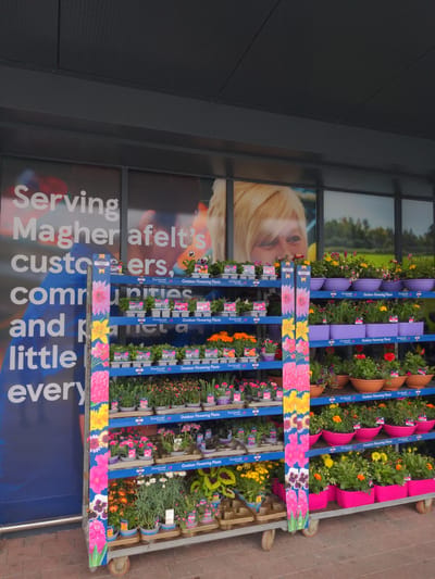 The image shows a display of outdoor flowering plants for sale outside a shop in Magherafelt, United Kingdom. The display is on wheeled shelving units, with various plants in pots and trays. The plants are of different colors and varieties, including pansies, dahlias, and lilies. The shop's signage is visible in the background, with the text "Serving Magherafelt's customers, communities and a little bit of everything." The overall scene suggests a retail environment focused on gardening and outdoor living.