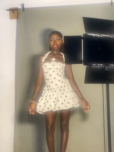 A young Black woman with dark hair pulled back poses against a plain grey backdrop, likely in a studio setting, given the presence of professional lighting equipment visible at the top of the frame. She is wearing a short, sleeveless white dress adorned with navy blue polka dots. The dress has a ruffled hem and a fitted bodice. She is accessorized with a delicate necklace featuring a floral pendant and a bracelet on her right wrist. Her makeup includes bold eyeshadow and lipstick. Her gaze is directed towards the camera with a confident expression. Her right arm is extended slightly upward and to her right, while her left arm is bent at the elbow, with her hand resting near her chest. The image is oriented vertically. There is no visible text in the image. The scene suggests a fashion or portrait photoshoot.