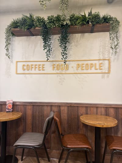 The image shows a section of a cafe interior. The left side of the frame features a white wall with a vertical wooden beam adorned with various green plants. To the right of this arrangement is a gold-framed sign with the words "COFFEE - FOOD - PEOPLE" written vertically. The right side of the image is dominated by a wood-paneled wall with several stools and chairs arranged around it. One stool has a light brown wooden seat, while the chairs have brown leather seats with black metal frames. A menu card is also visible near the bottom right corner of the image. The cafe's specific location in Mole Valley, United Kingdom cannot be determined from the image.