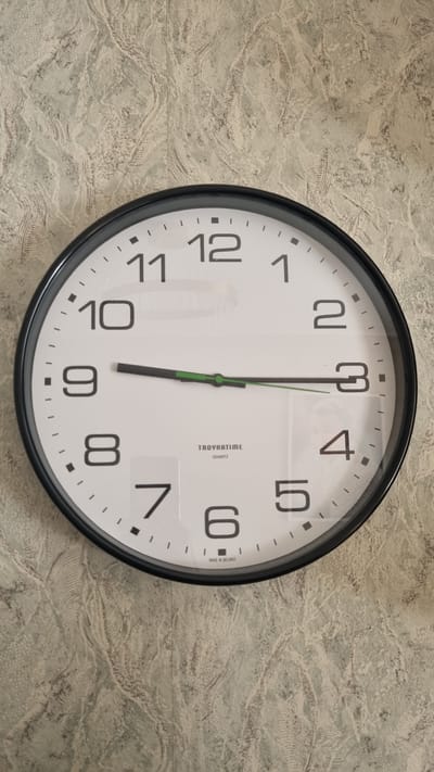The image shows a round analog clock with a black frame hanging on a textured wallpaper. The clock face is white with black Arabic numerals. The hour hand is pointing to the 9, and the minute hand is pointing to the 3. The second hand is green. The brand name "TAOYHATIME QUARTZ" and "MADE IN BELARUS" are visible at the bottom of the clock face. The background wallpaper has a marbled pattern in shades of gray and beige. There are no people or animals present in the image. The lighting suggests an indoor setting, possibly a room in a house or office. The clock is positioned centrally in the frame.
