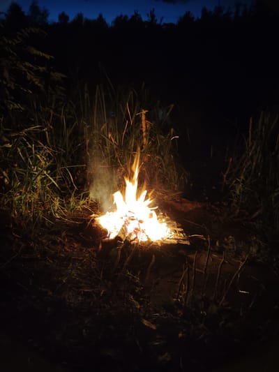 In Balyko-Shchuchynka, Ukraine, a campfire burns at night in a rural setting. The flames are bright orange and yellow, with smoke rising above them. The surrounding area is dark, with tall grass and foliage visible. A line of trees forms a dark silhouette against the deep blue night sky in the background. The ground appears to be covered in dirt and dry vegetation. There are no people visible in the image.