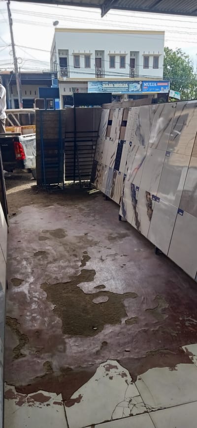 The image captures a scene outside a building in Meunasahlhok, Indonesia, likely a commercial establishment selling building materials. In the foreground, the ground appears to be a cracked and weathered concrete or tiled surface, with large patches of material peeling away, revealing a darker underlying layer. Various tiles, likely for flooring or walls, are displayed upright on stands to the right of the image. These tiles exhibit a range of patterns, including marble-like veining and geometric designs.
In the middle ground, a black pickup truck is parked, with its rear visible. To the left of the truck, what appear to be stacks of boxes or construction materials are partially obscured. A person in a white shirt is partially visible near the truck. Behind the truck and materials, a multi-story building with a white facade and visible windows stands under a cloudy sky. A sign with blue and white lettering, reading "TOKO RAJA MULIA ELEKTRONIK," is affixed to the building, indicating it is a store. The scene suggests an outdoor storage or display area associated with a business.